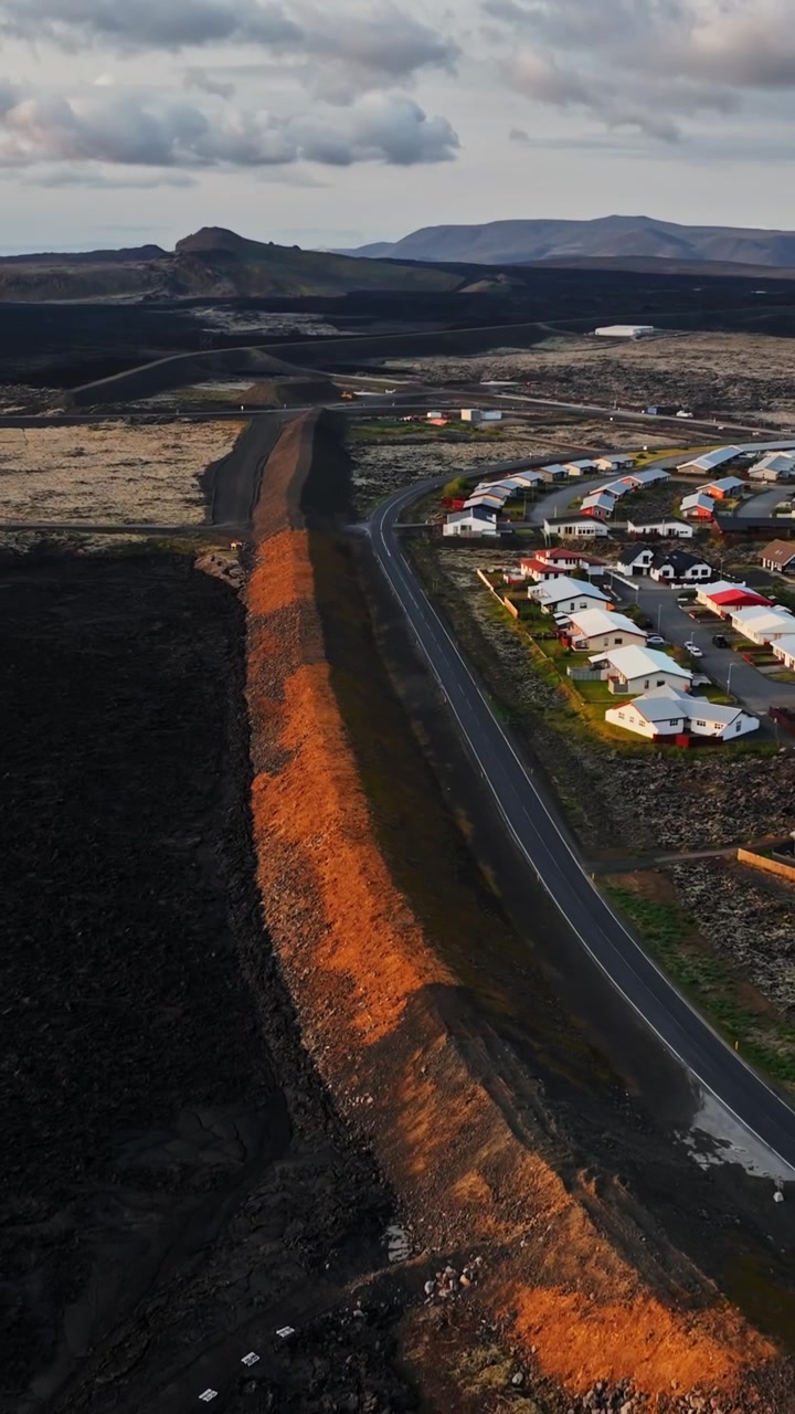Vista aerea verticale da drone lungo l'argine di difesa dalla lava di Grindavik, che mostra il terreno di eruzione nero su un lato e una strada, case e paesaggio vulcanico sull'altro.