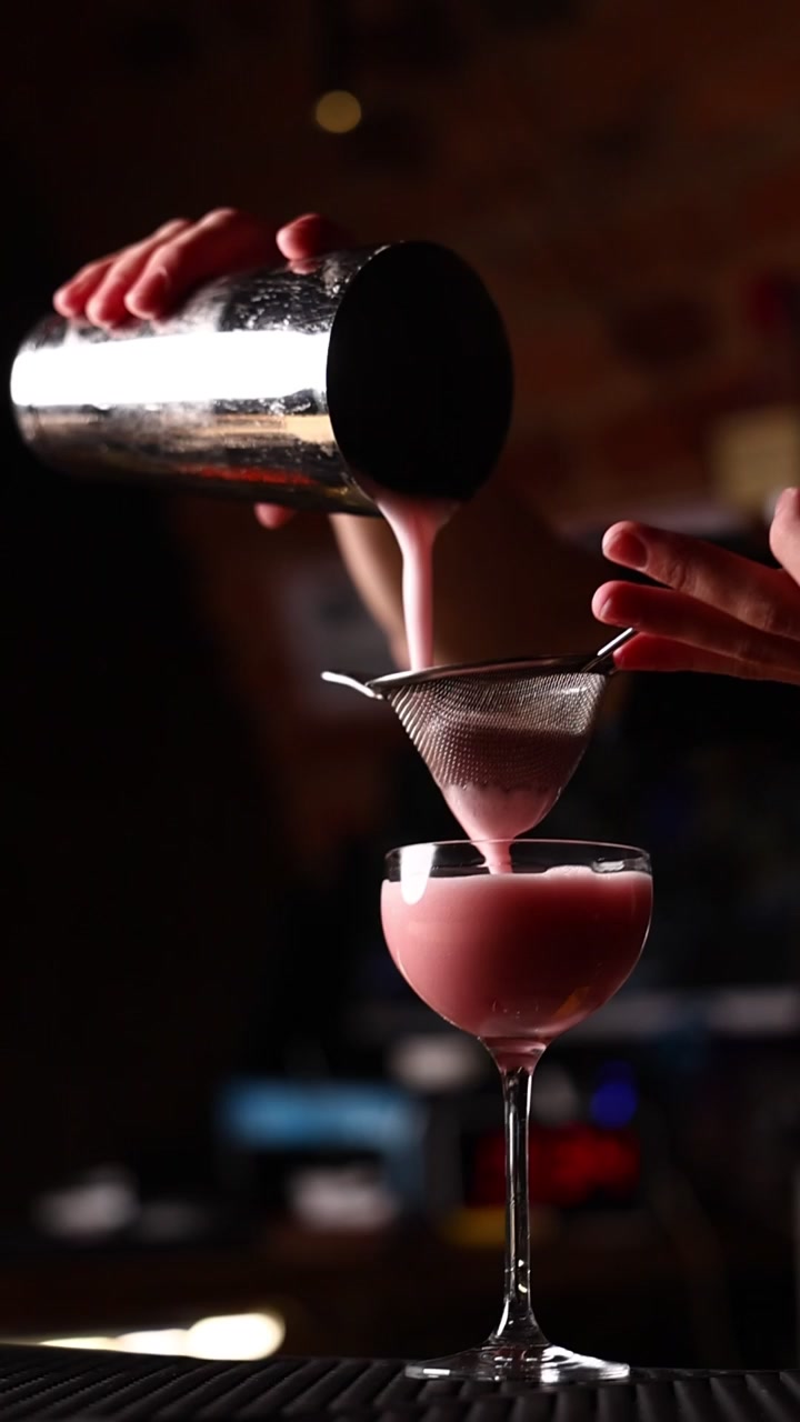 Bartender, der sier en lyserød cocktail ned i et glas i en bar
