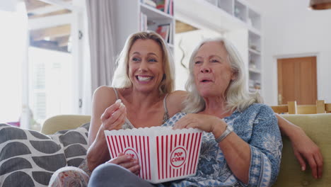 madre con hija adulta comiendo palomitas de maíz viendo una película en el sofá en casa juntos