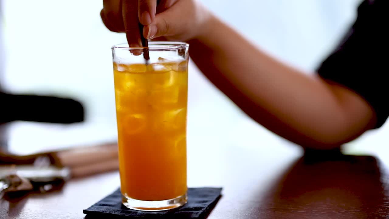 Hand stirs iced orange juice in glass, natural light, wooden table, casual dining atmosphere