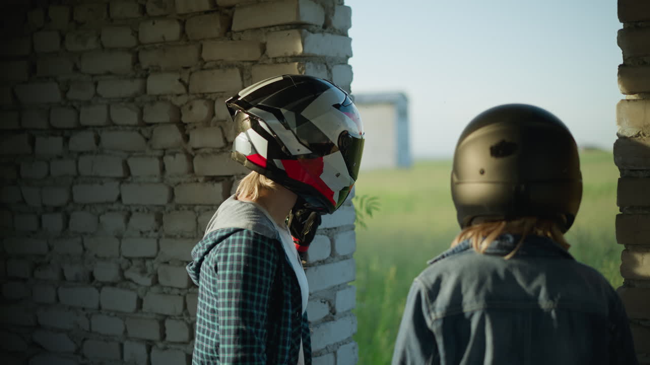 vista lateral de dos hermanas con cascos de pie dentro de un edificio abandonado, mirando a un vasto campo verde a través de una abertura, con un objeto visible en el fondo