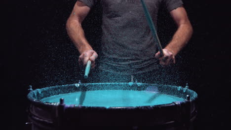 Man-playing-a-drum-that-is-filled-with-blue-paint-that-splatters-when-the-drumsticks-hit