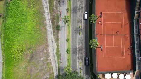 Smooth-car-overhead-tracking-by-a-drone-next-to-tennis-club-courts,-bicycle-riders,-palms,-trees,-and-gardens