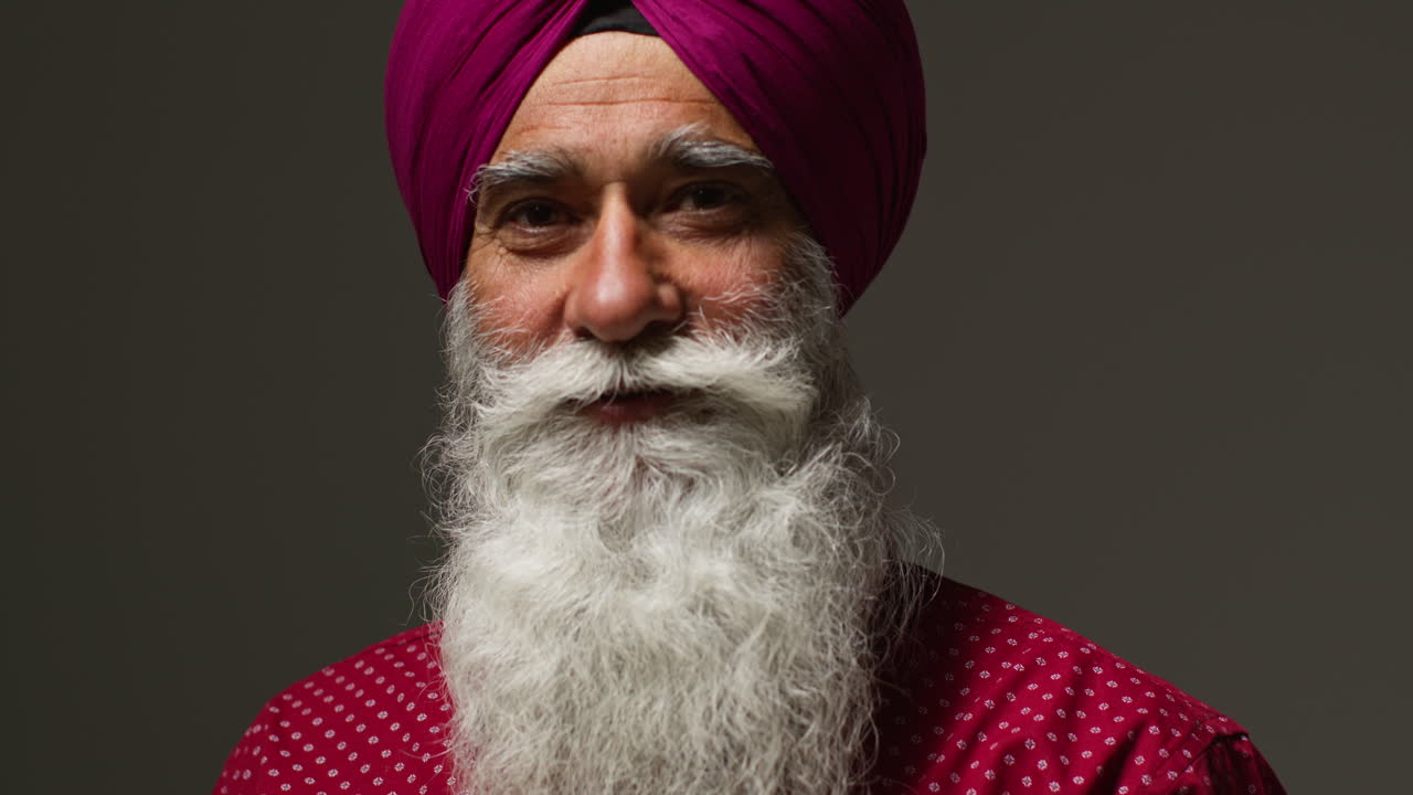 Close Up Low Key Studio Lighting Portrait Of Senior Sikh Man With Beard Using Salai Needle When Putting On Turban Against Dark Background