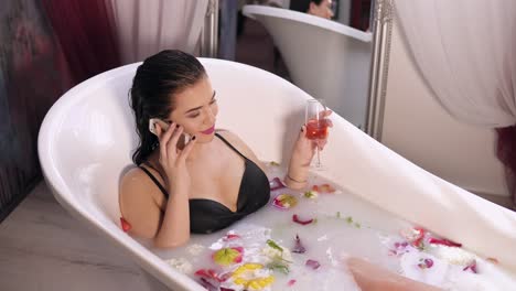 Beautiful-young-woman-talking-on-the-phone,-relaxing-in-milk-bath-and-drinking-sparkling-wine-from-a-glass.-Shot-in-4k