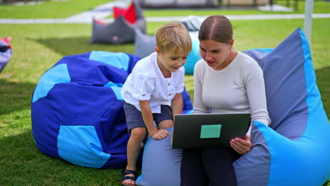 blonde woman and her son resting in bean bag chairs watching video on laptop. family resting outdoors in summer. blurred backdrop.