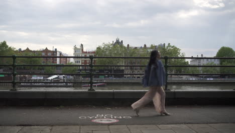 fashionable latina tourist with black wavy hair and a jean jacket walking and spinning in london with a view of boats and buildings behind her