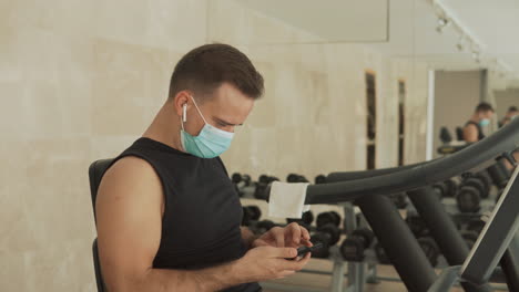 young strong man with face mask uses a smartphone and an exercise machine in the gym