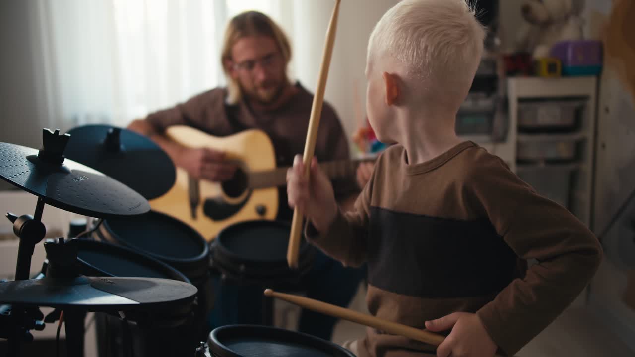 A happy albino boy with white hair plays on a special electronic drum kit using yellow drum sticks and his father, a blond man with long hair with a beard and glasses, plays the guitar with his son