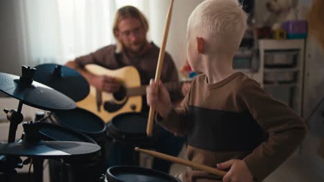 Ein-Glücklicher-Albino-Junge-Mit-Weißem-Haar-Spielt-Auf-Einem-Speziellen-Elektronischen-Schlagzeug-Mit-Gelben-Trommelstöcken-Und-Sein-Vater,-Ein-Blonder-Mann-Mit-Langen-Haaren,-Bart-Und-Brille,-Spielt-Mit-Seinem-Sohn-Gitarre