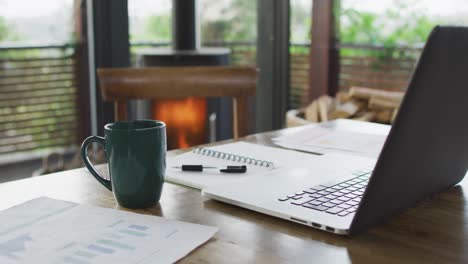 Escritorio-De-Trabajo-En-Casa-Con-Computadora-Portátil,-Libreta-Y-Taza-De-Café-En-La-Sala-De-Estar,-Con-Fuego-En-Una-Estufa-De-Leña