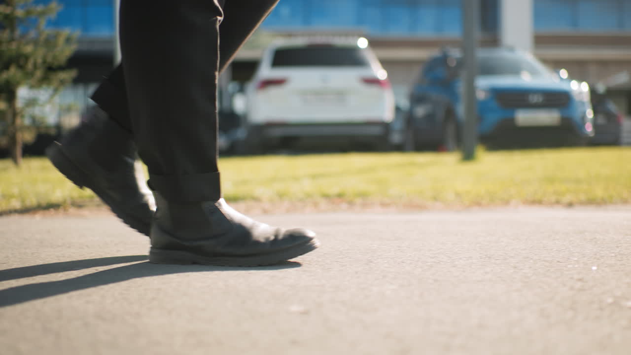 빛이 있는 날, 지상에 그늘이 떨어지고, 주차된 자동차, 초록 잔디, 현대 도시 건물 등이 배경으로, 타르로 된 도로에서 자신감 있게 고 있는 사람의 측면 풍경