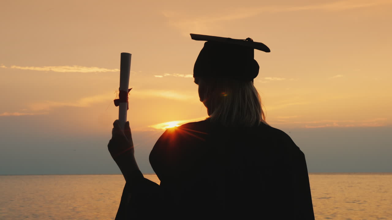 un soltero con un diploma en la mano y una gorra de graduado mira el amanecer sobre el mar