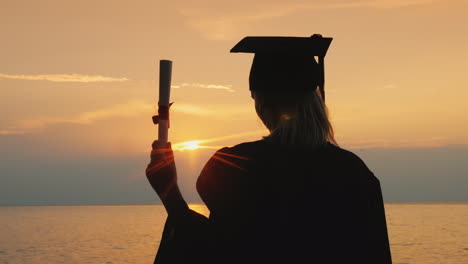 a bachelor with a diploma in hand and a cap of a graduate looks at the sunrise over the sea