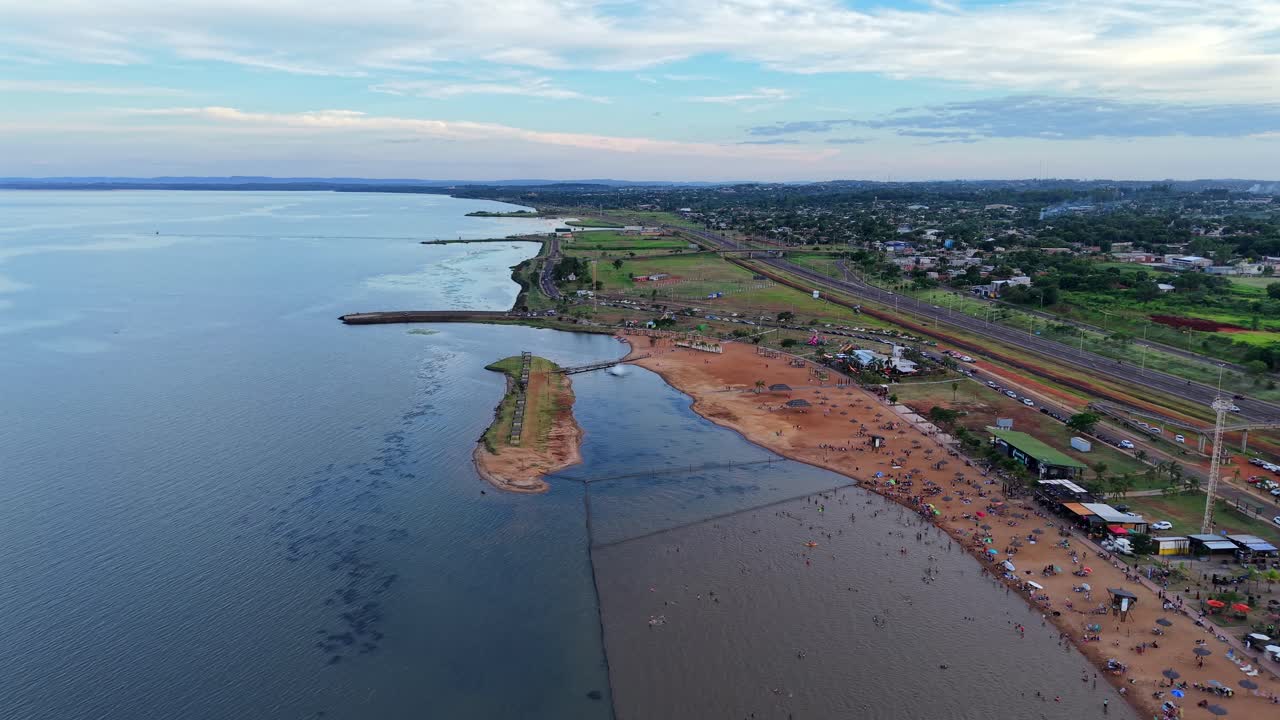 Il Balneario Costa Sur a Posadas, Misiones, Argentina, mostra una spiaggia sabbiosa, un lungo litorale, un molo e una barriera frangiflutti, palme paesaggistiche e folle che si godono il fiume Paraná al tramonto, con rivelazione lenta