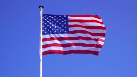 An-American-flag-flying-in-the-wind-against-a-blue-sky-backdrop,-slow-motion