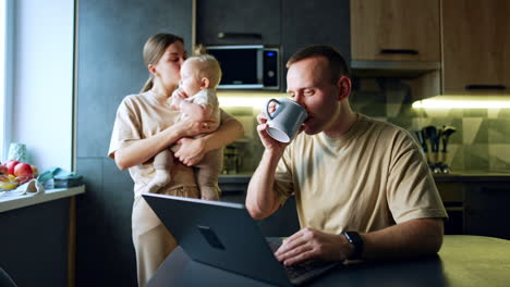 man sitting in front of the laptop taking a cup and drinking. woman with baby in hands stands behind.