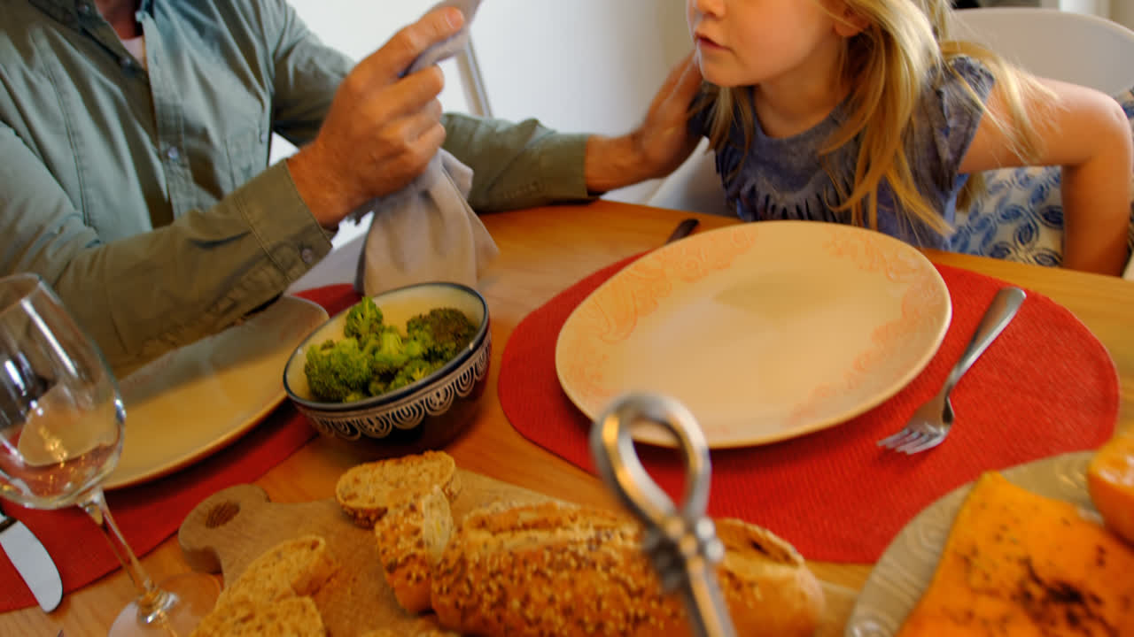 vista frontal de un padre caucásico de mediana edad está limpiando la boca de su hija en la mesa del comedor en casa 4k