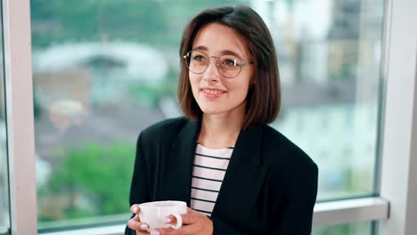 happy resilient caucasian brunette wearing glasses stands at window. positive woman portrait with coffee cup in hands.