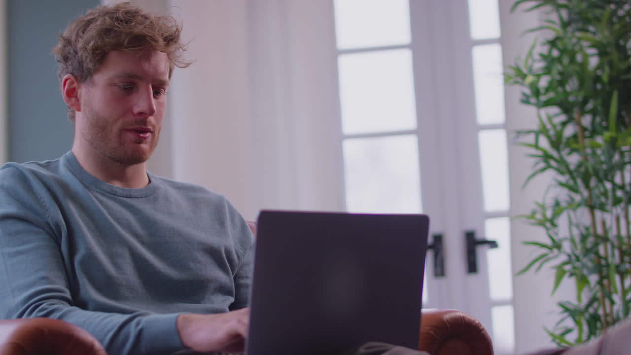 Young Man Sitting In Chair At Home And Working On Laptop