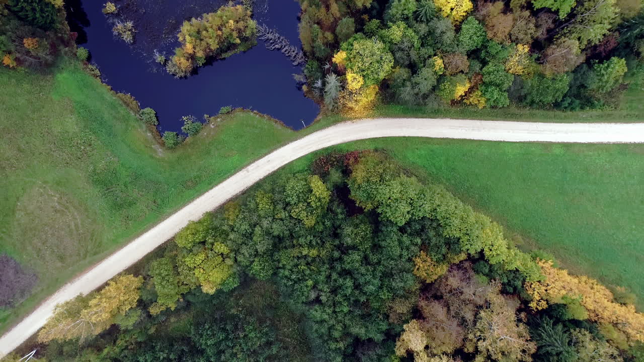 Aerial Bird's eye view over lake surrounded by colorful autumn forest beside a narrow pathway on a countryside at daytime