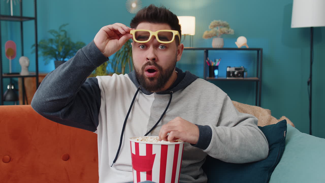 Young man sits on couch eating popcorn and watching interesting tv serial sport game online at home