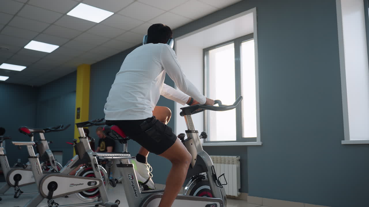 Boy in white long-sleeve top and black shorts pedals on stationary bike in gym, with multiple identical bikes lined up beside him under bright ceiling lights and large windows