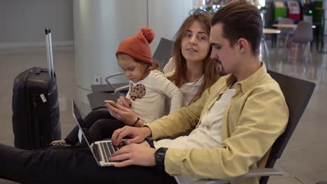 young father working on laptop on airport with his family while waiting for departure. long haired mother entertains her little