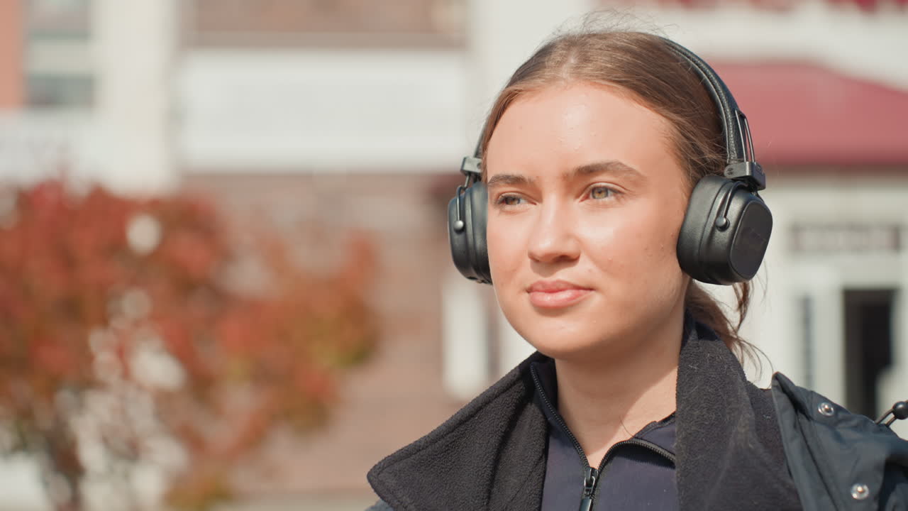 Junge Frau, die im Sonnenlicht Kaffee trinkt, mit Kopfhörern und Nahaufnahmen ihres Profils, gelassener Gesichtsausdruck, geringe Schärfentiefe Bokeh, Coffee-to-go-Becher und strukturierte Jacke, Kleinstadtstraße und Schaufenster