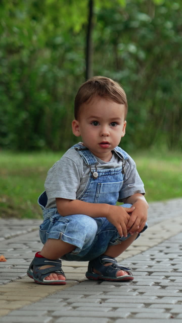 Beautiful Caucasian kid sits squatted on the paved road. Kid looks thoughtfully ahead and then runs. Vertical video