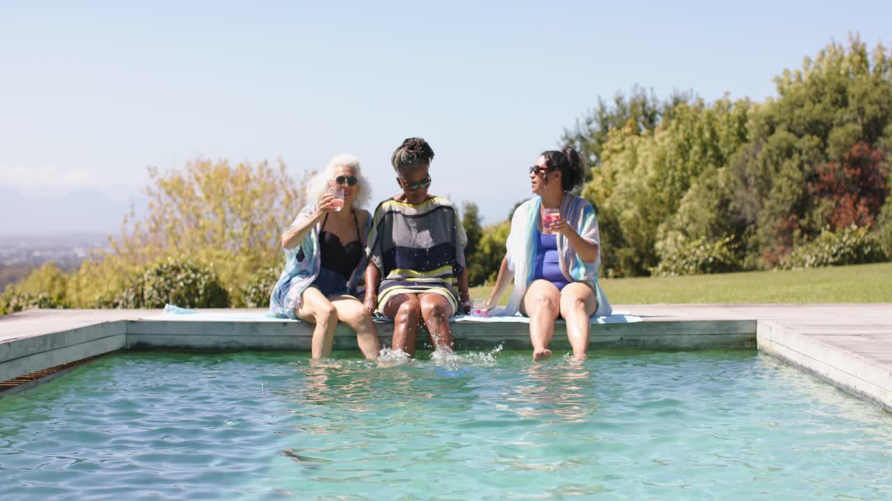 Left woman raising cup, group splashing feet and toasting at poolside with pink drinks in swimsuits