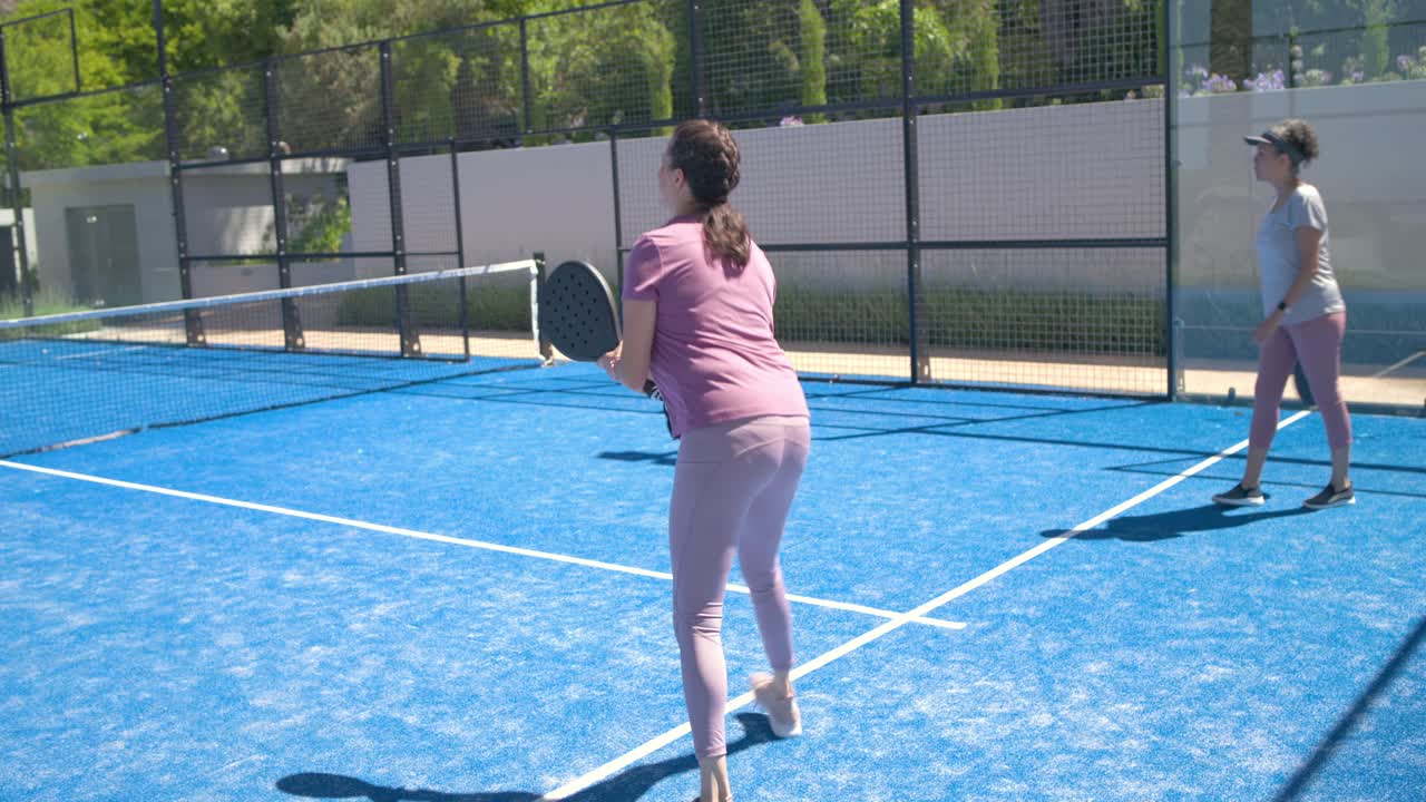 Mulheres diversas praticando um rali em quadra azul com raquetes, uma parceira lançando a bola, com a jogadora de rosa rebatendo.