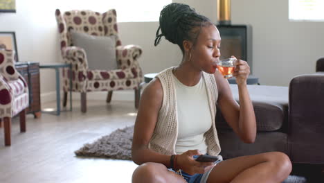 african american young woman sitting, sipping tea, holding smartphone, at home