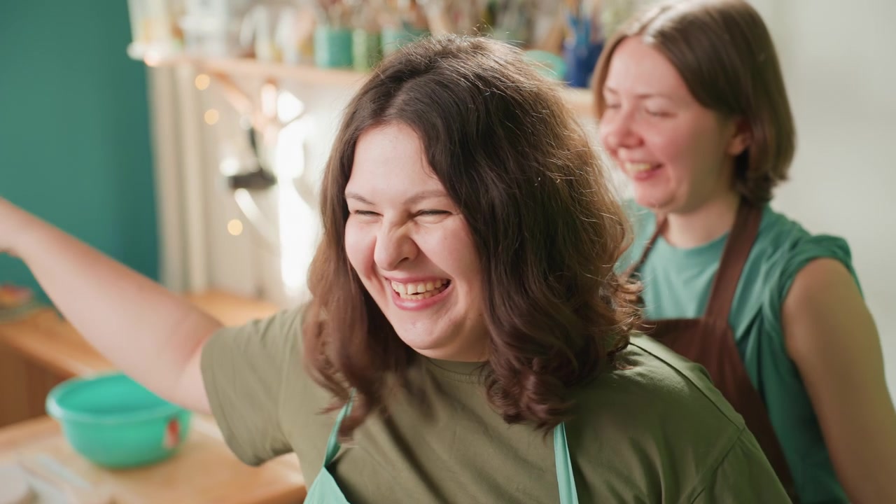 Two caucasian potters laughing and cheering in studio, apron and tools visible, shelves of glazes and bowls, playful high spirits, teamwork and learning, candid joyful moment after successful piece