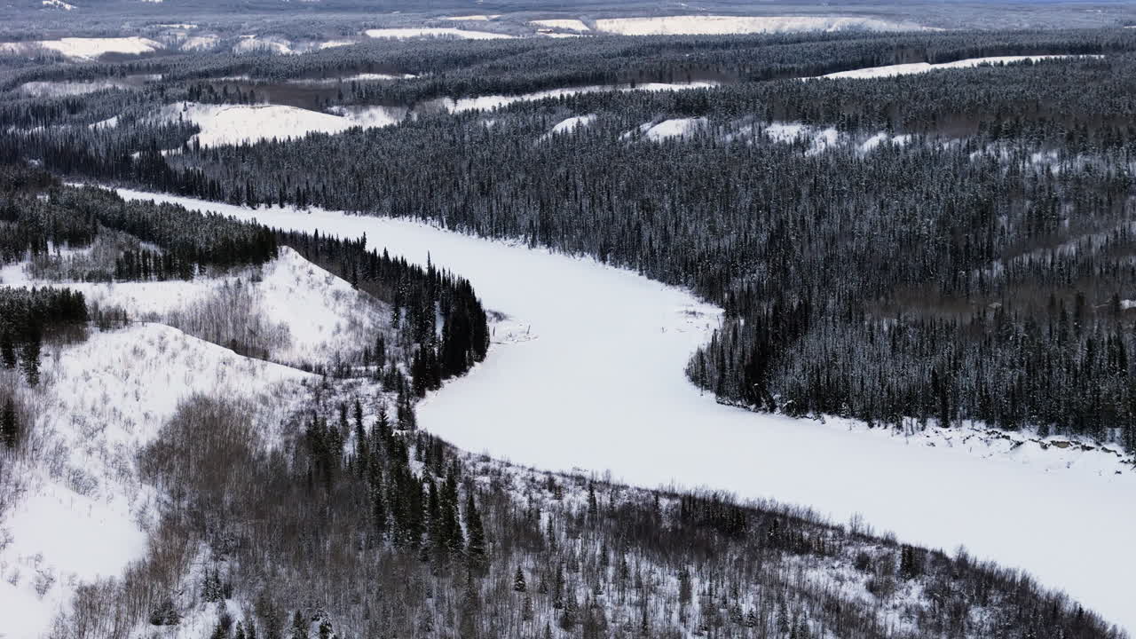 A sweeping aerial shot captures the Takhini River carving through a snowy Ibex Valley, framed by fir-lined hills. The vast Yukon winter landscape evokes a sense of isolation and beauty.