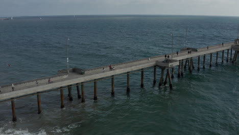 AERIAL-flight-next-to-pier-at-Venice-Beach,-Los-Angeles-California-in-cloudy-weather-with-waves-in-ocean