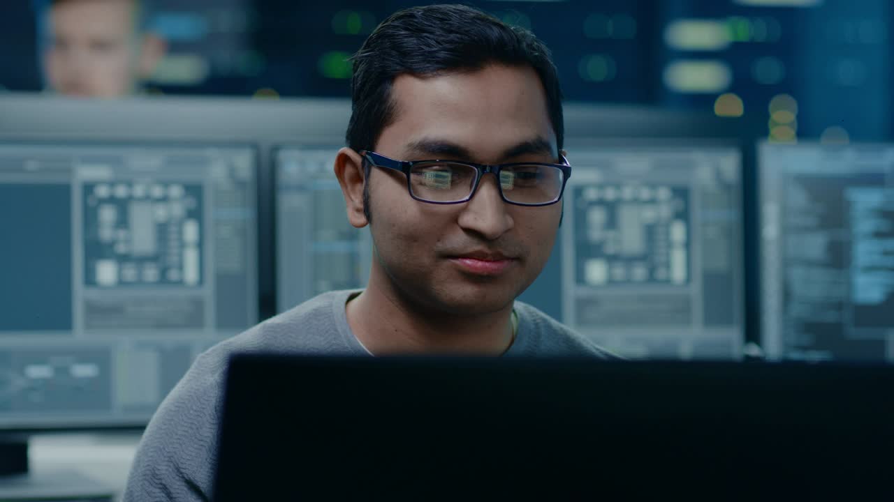 Portrait of a Smart and Handsome Software Developer Wearing Glasses Working on a Personal Computer, Smiles at the Camera. In the Background Unfocused Personal Computers with Screens in Technical Room
