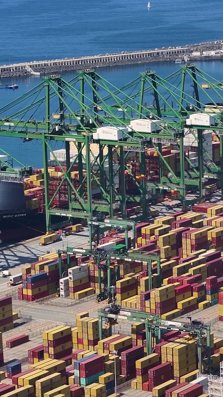 A container terminal with numerous shipping containers stacked and organized at a busy cargo port. Large cranes are actively loading and unloading vessels.