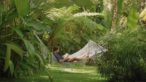 young man lying in hammock reading book in jungle paradise surrounded by plants