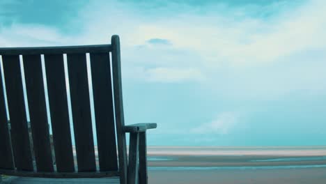 a single, wooden beach chair sits facing a sunny beach at low tide where veins of water stretch towards the open waters under an angry sky brewing up dark, rainy clouds on the horizon