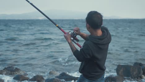 young man struggling to real in fishing rod at windy seaside coastline