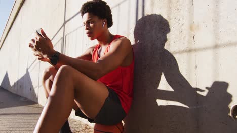 Happy-african-american-man-in-city,-using-smartphone-with-wireless-earphones,-sitting-on-basketball