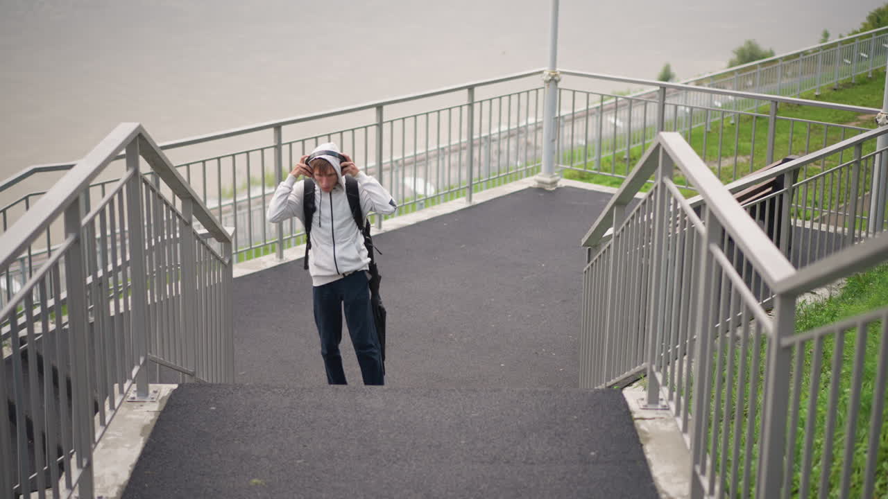 흐린 하늘 아래 강변 계단을 올라가는 후드티와 백팩을 착용한 남자가 접힌 우산을 들고, 잠시 멈춰 후드를 고쳐 쓰고 물가를 바라보는 모습, 금속 난간과 젖은 아스팔트, 우울한 통근자의 모습