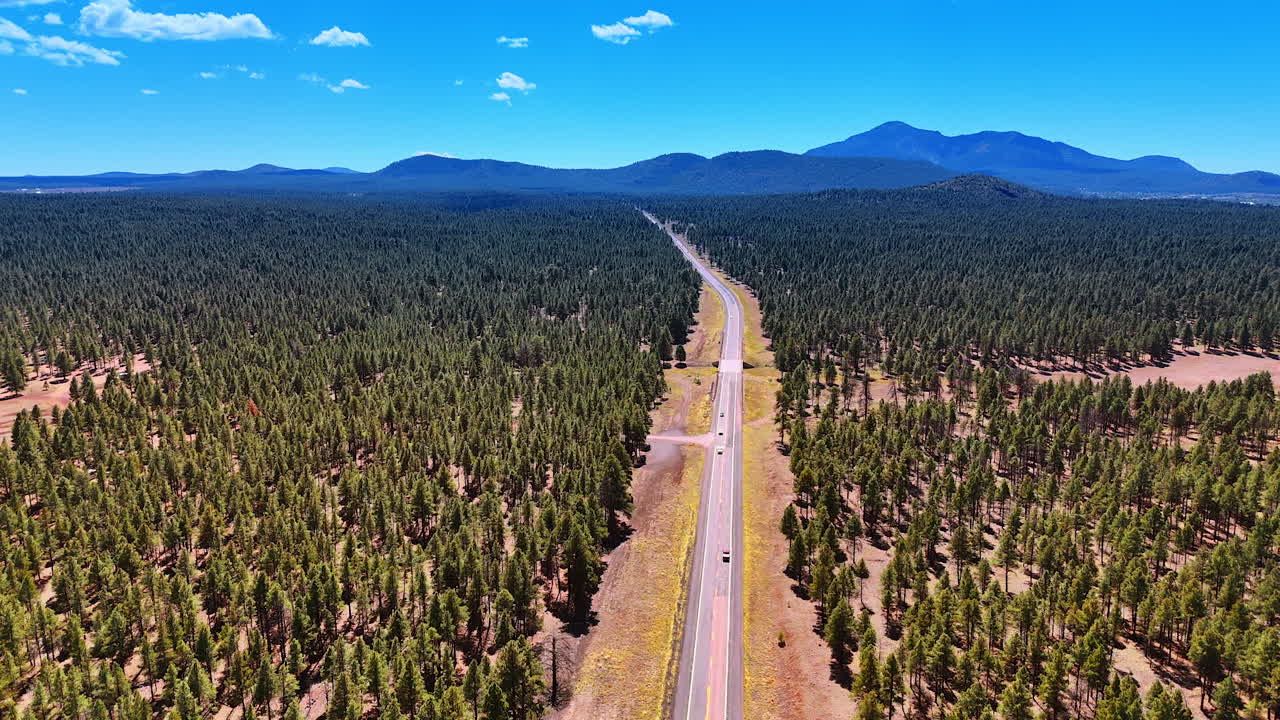 Few cars move by the highway in the pine tree wood covering the vast valley. Hazy mountain silhouettes at backdrop. Arizona, USA