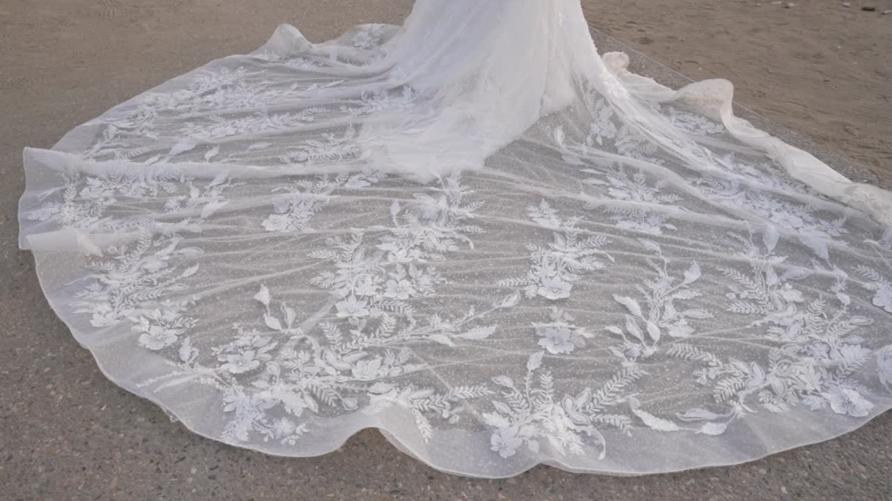 A bridal gown features intricate lace patterns spreading out on the ground. The outdoor setting shows natural light highlighting the dress. The fabric lies gently on the surface