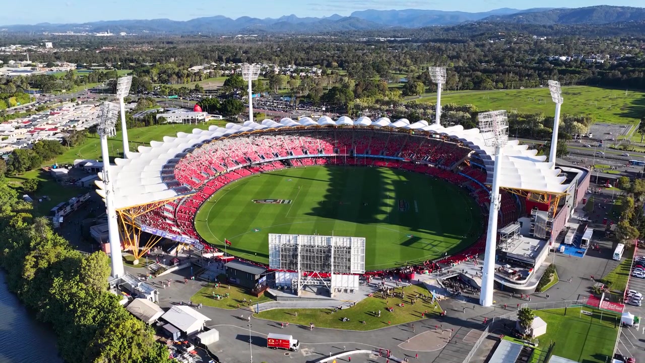 High-angle drone shot captures a circular sports arena with green field under bright sunlight.