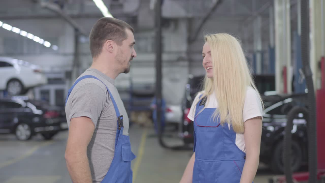 молодой кавказский мужчина и женщина показывают жест силы, смотрят в камеру и улыбаются. профессиональные автомеханики в рабочих халатах стоят на рабочем месте в ремонтной мастерской. профессия, профессия, работа.