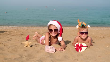 children lie on the beach wearing a santa hat and antlers. little boy and girl are looking at the camera and waving hello. merry christmas. winter vacation. the sea is in the background. copy space