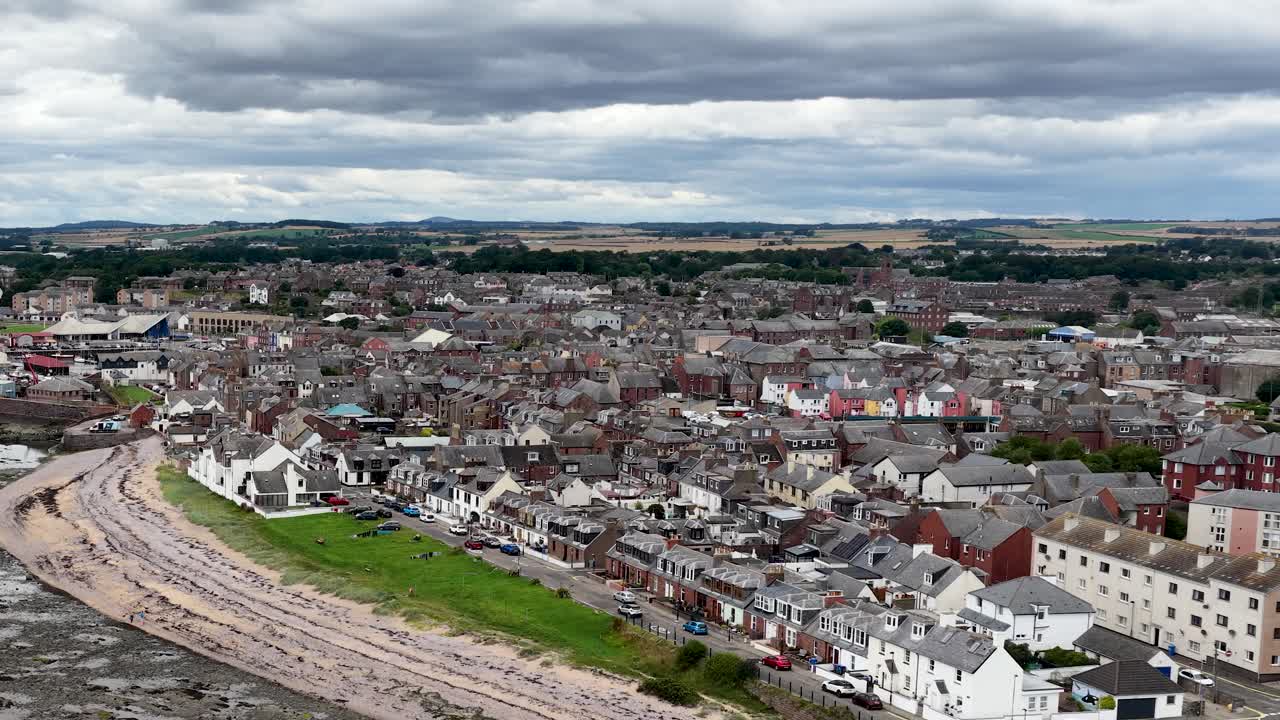Drone pans over Dundee coastal town, harbor, rocky shoreline, and green fields under clouds