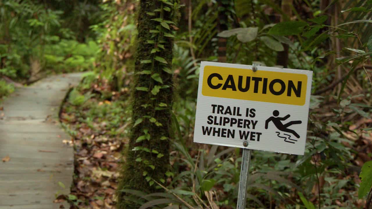 A yellow caution sign stands beside a wooden boardwalk path through dense tropical jungle foliage